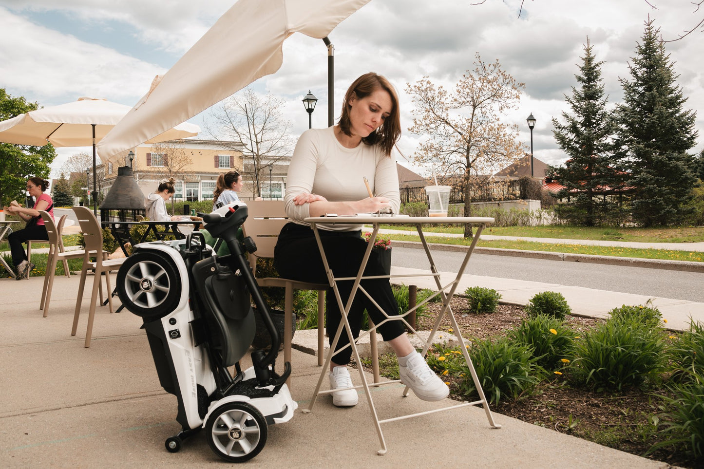 Woman seated at an outdoor cafe table beside a folded Heartway Transit 4 scooter
