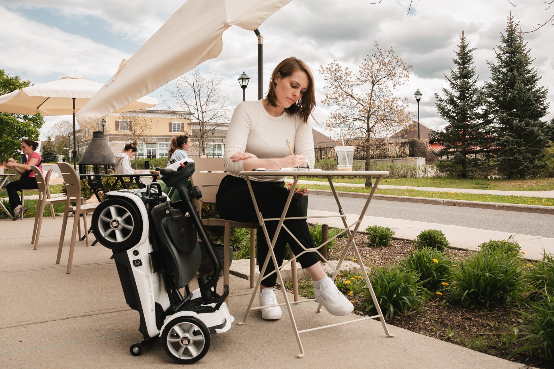 Woman seated at an outdoor cafe table beside a folded Heartway Transit 4 scooter