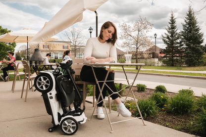 Woman seated at an outdoor cafe table beside a folded Heartway Transit 4 scooter